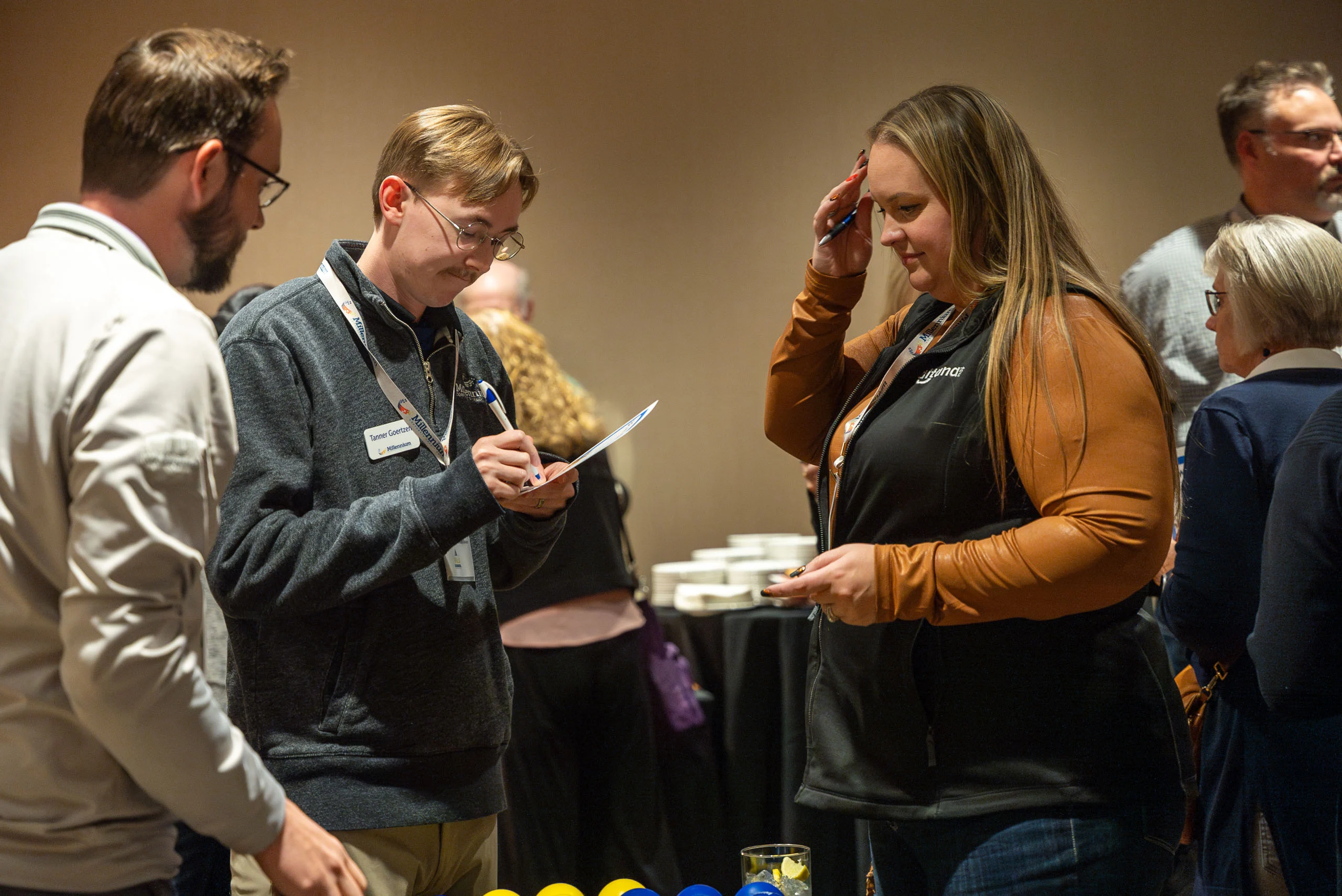 Two attendees sign papers at a registration table while a woman stands nearby at a conference; cups and lemon slices are visible at the front of the table.