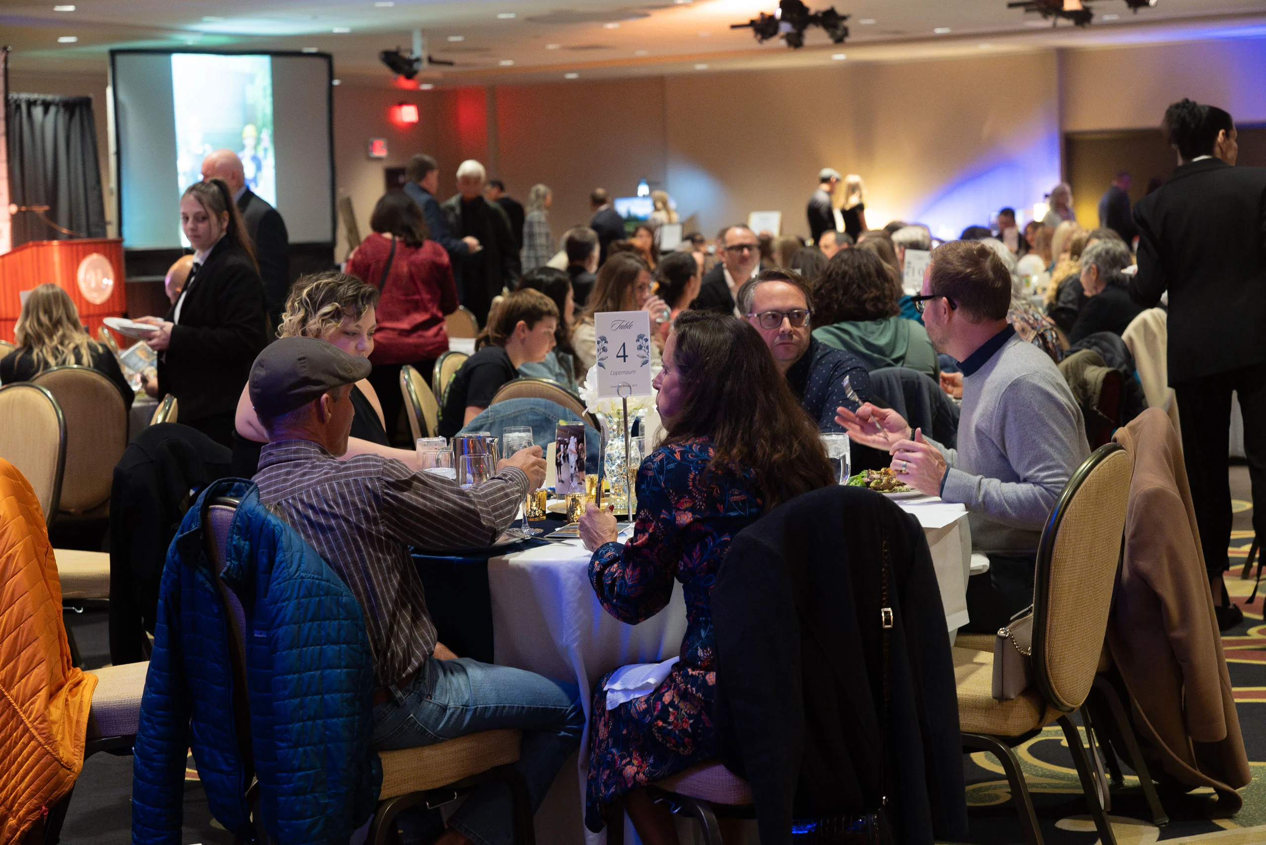 Bustling banquet scene with guests seated at round tables, eating and talking; a 'Table 4' sign visible in foreground.