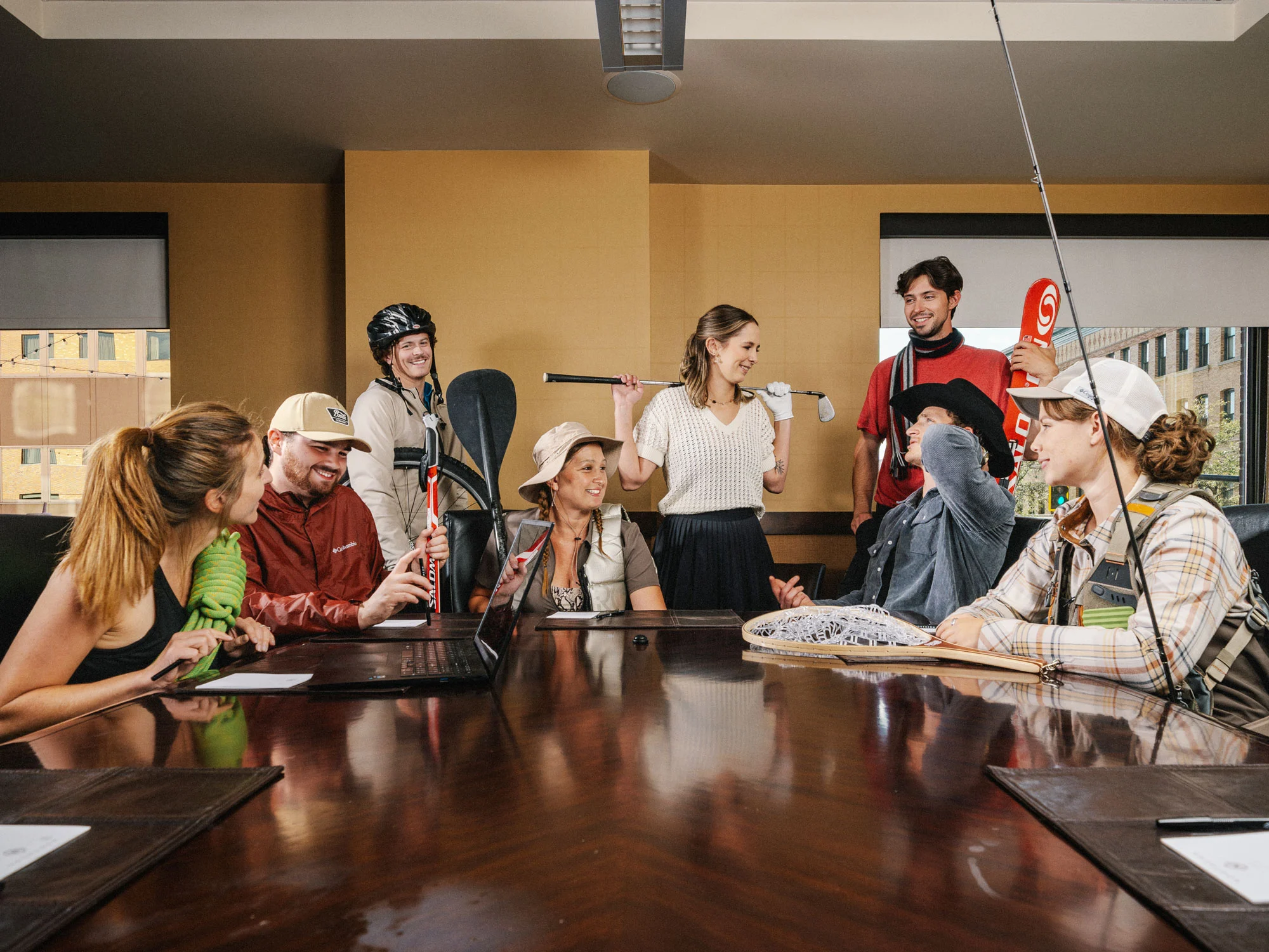 Group of people around a conference table, unpacking sports gear like skis and helmets in a casual meeting setting inside a room.